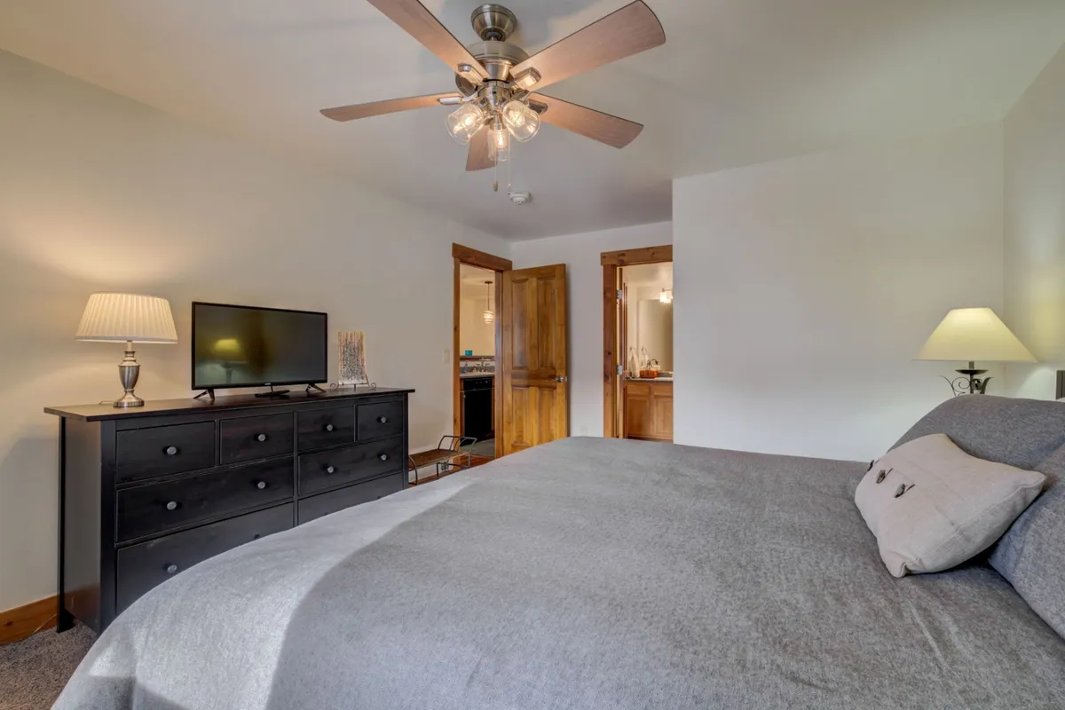 A serene bedroom is shown with a bed featuring gray bedding, a black dresser with a television, and—most notably—a ceiling fan with a light fixture overhead. Twin nightstands with lamps flank the bed, enhancing the room’s restful mood.