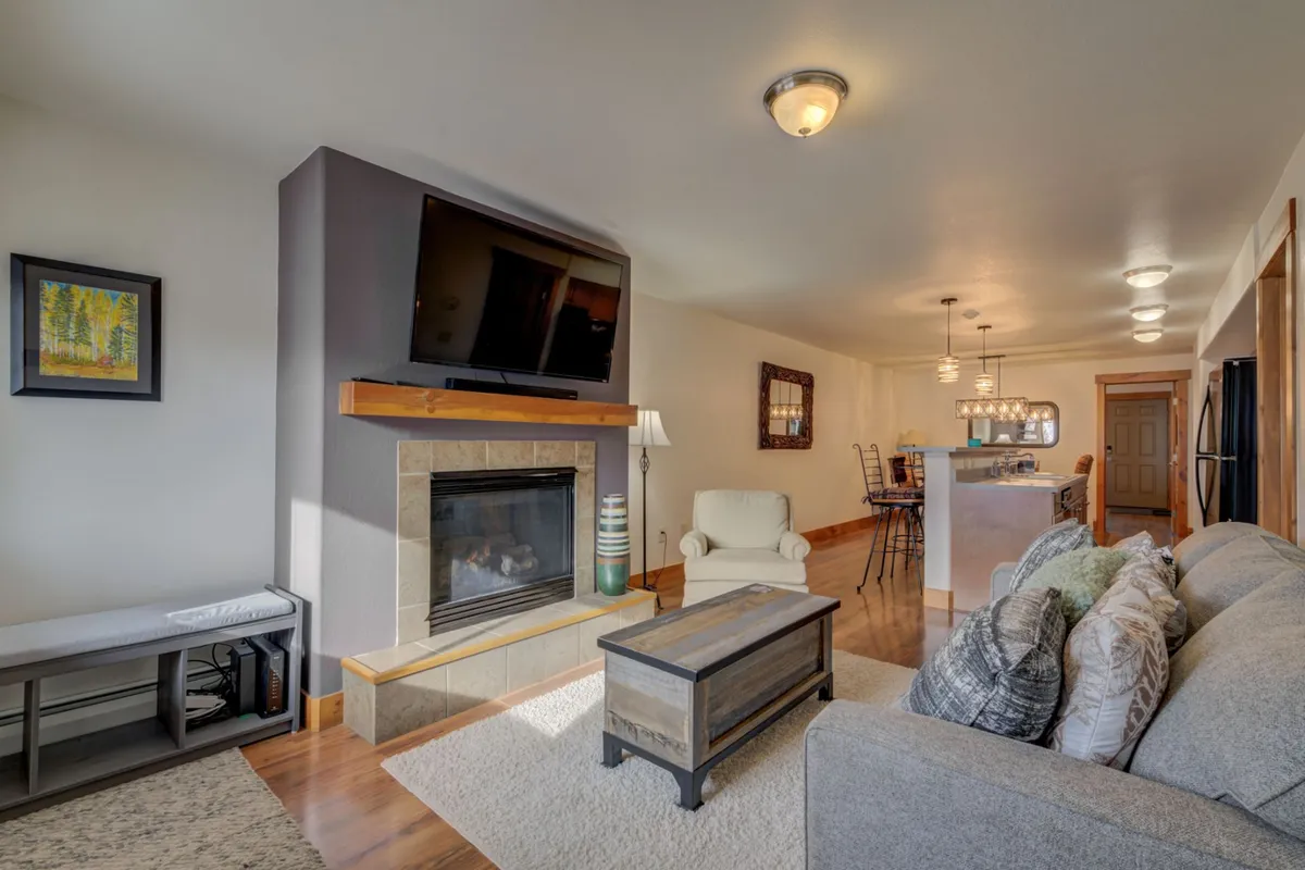 A wide-angle view of the open-plan living space, showcasing a cozy sofa facing a stone gas fireplace, a flat-screen TV above the mantle, and a spacious dining area adjacent to a modern kitchen. The room is warmly lit and decorated with a colorful painting, creating an inviting atmosphere for gatherings and relaxation.