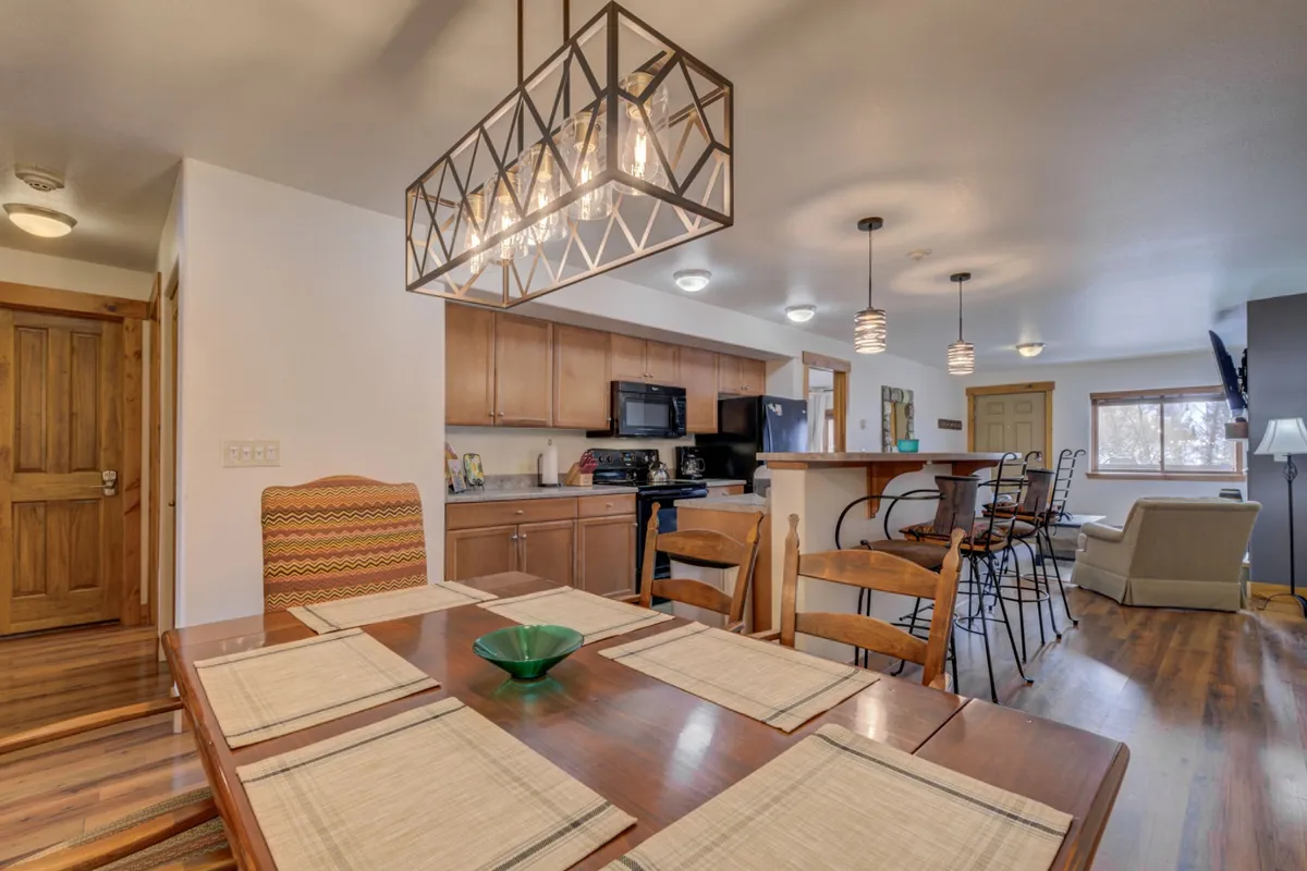 A wide view of a welcoming dining space with a dining table, chandelier, kitchen island, sofa, and a bright window. The area feels communal and versatile, ideal for groups returning from outdoor excursions or preparing for a day of activities.