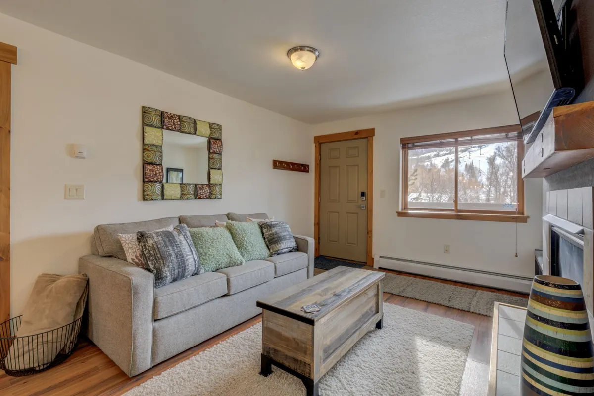 An inviting living area with a sofa, decorative pillows, a wooden coffee table, a mirror, and a window that lets in natural light. The entry door is visible, reinforcing the welcoming atmosphere as you step into your private haven.