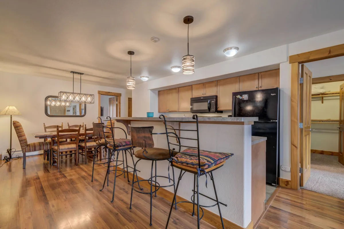 A bright, welcoming kitchen and dining area featuring a large bar counter with stools, a wooden dining table set for several guests, and stylish lighting fixtures. The kitchen is modern and open, inviting for both casual breakfasts and celebratory dinners.