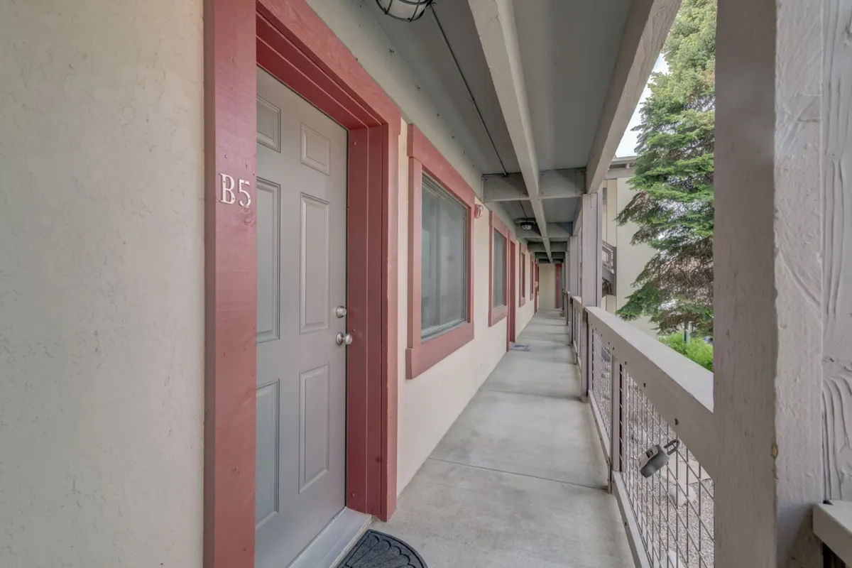 A view down a bright interior corridor with windows, a visible door, and greenery outside, conveying a sense of arrival and seamless transition from the condo to the outdoors and community spaces.