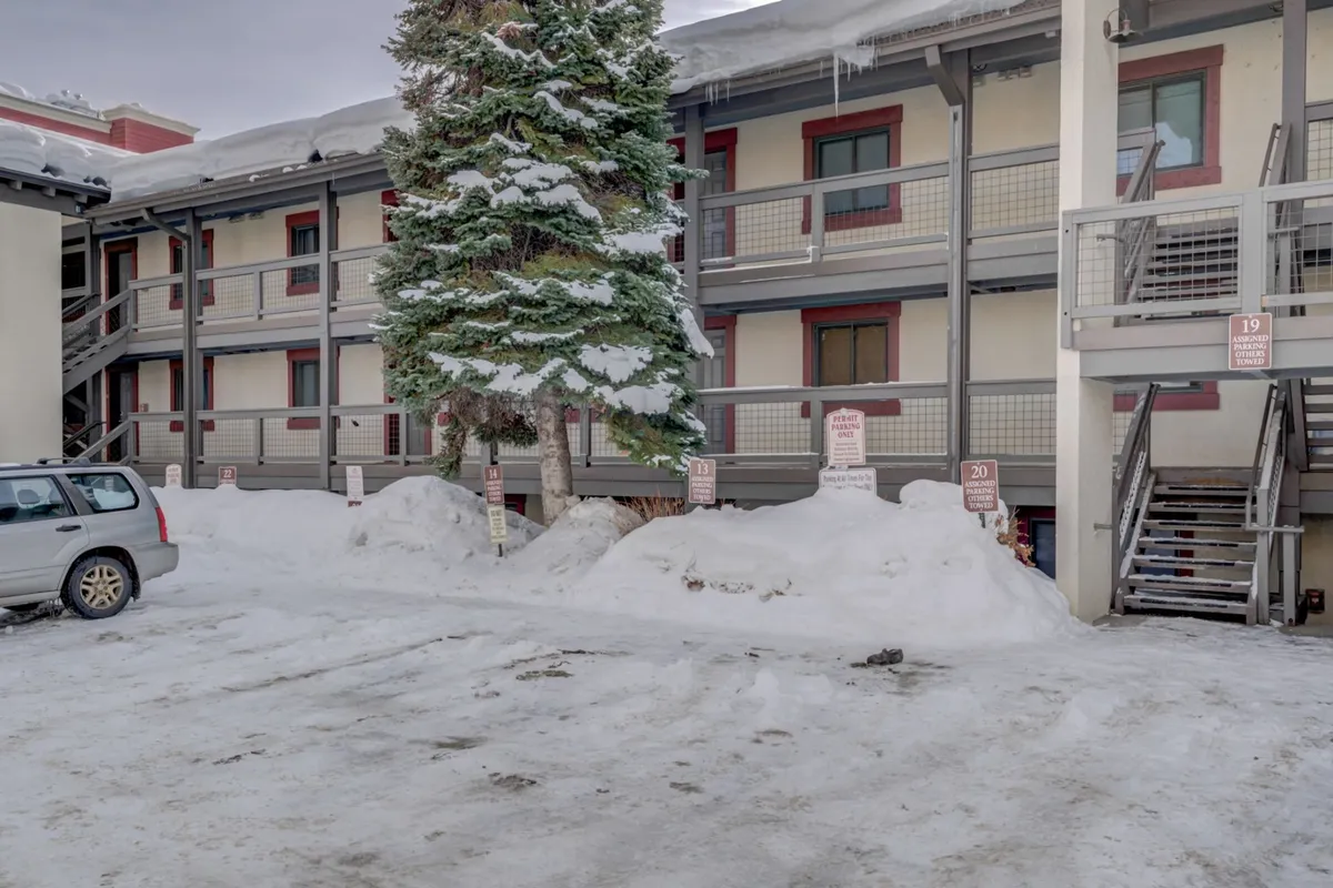 This exterior shot features the building's facade, adjacent pine trees, and the parking area blanketed in snow. The tranquil winter scene hints at the property's proximity to downtown attractions, suggesting a peaceful retreat that remains connected to the energy and convenience of Steamboat Springs.