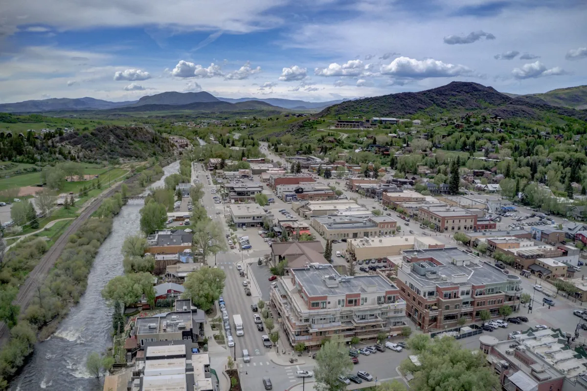This vibrant exterior shot captures the lively town center with the Yampa River flowing nearby, lush greenery, mountains in the background, and people walking, shopping, and dining. The energetic atmosphere showcases the condo’s proximity to both urban amenities and natural beauty.