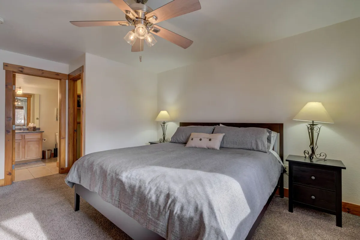 A tranquil bedroom featuring a king-sized bed with plush bedding, flanked by nightstands with lamps and a ceiling fan overhead. The soft light from outside gives the room a serene, restful vibe, and the overall decor is understated yet inviting.