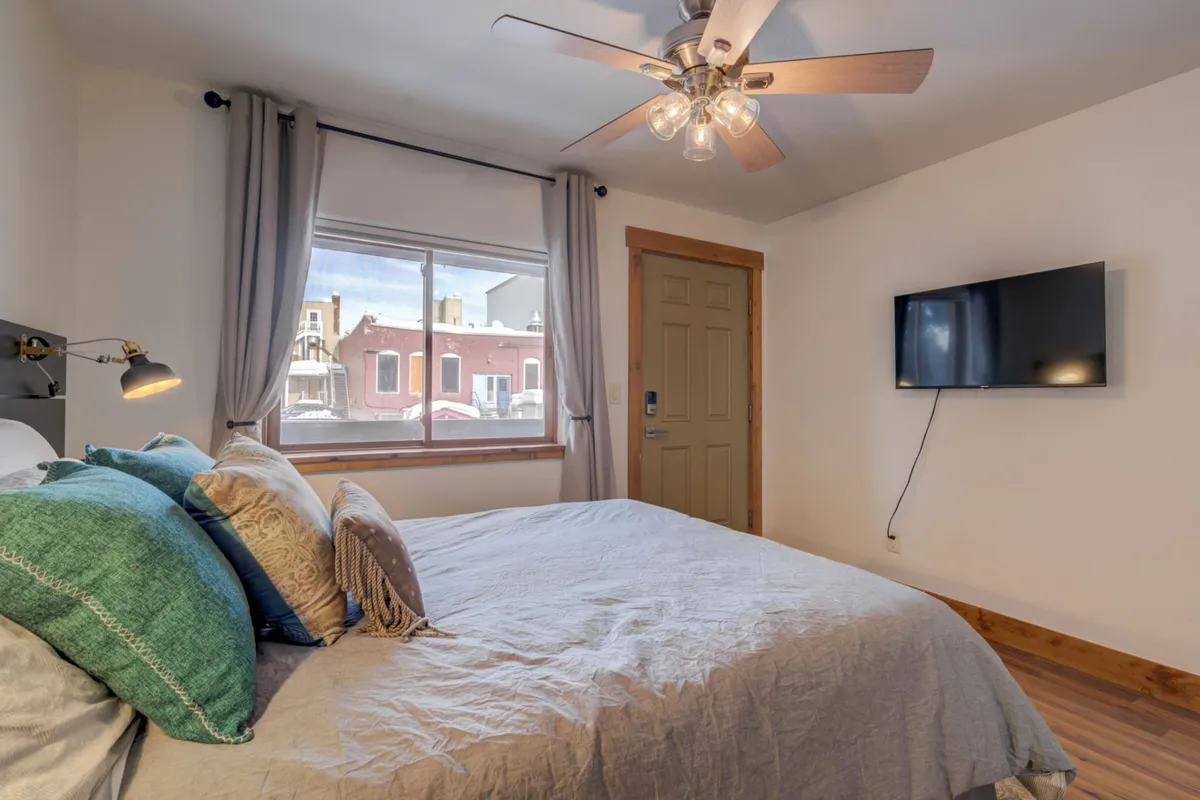 A cozy bedroom with decorative pillows on the bed, a window letting in natural light, a ceiling fan, and a television mounted on the wall. The space feels inviting and is designed for comfort, offering both a relaxing environment and entertainment options.