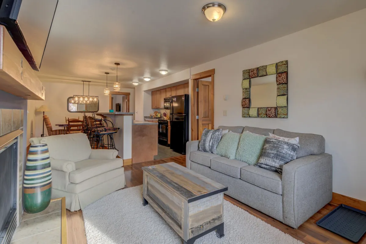 This bright and welcoming living area features a plush sofa adorned with decorative pillows, a wooden coffee table, and a prominent fireplace. In the background, the open kitchen and dining table are visible, suggesting a seamless flow for relaxing and socializing after a day outdoors.