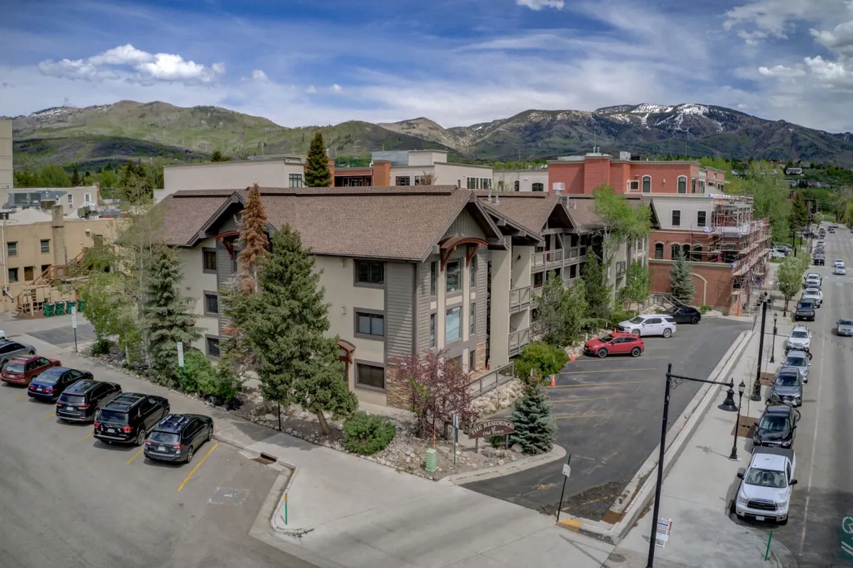 An exterior view of the building with a distinctive sloped roof, surrounded by lush trees and framed by distant mountains. The scene conveys a welcoming, serene atmosphere and hints at the proximity to parks, trails, and outdoor amenities. People are seen walking in the nearby park, exploring local shops, and enjoying outdoor dining, reinforcing the spirit of community and access to adventure.