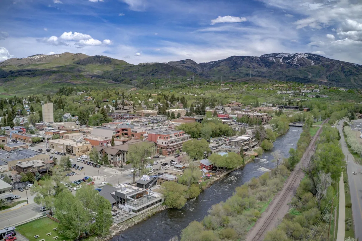 A vibrant riverfront scene featuring the Yampa River bordered by green trees, with town buildings nestled at the base of a scenic mountain backdrop. The setting is lively, suggesting opportunities for riverside activities such as walking, picnicking, and enjoying the natural beauty. The overall mood is energetic and welcoming, highlighting the river as a central feature of the community.