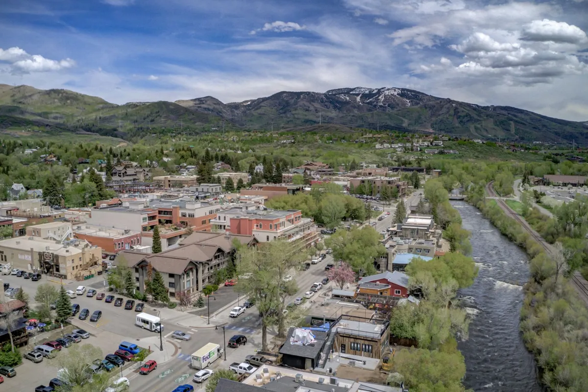 The image showcases a vibrant view of downtown Steamboat Springs with the Yampa River flowing in the foreground, tree-lined streets bustling with life, charming town buildings, and the majestic mountains rising in the background. People can be seen enjoying riverside walks, dining outdoors, and exploring local shops, all set against a scenic mountain backdrop that hints at endless outdoor adventures.