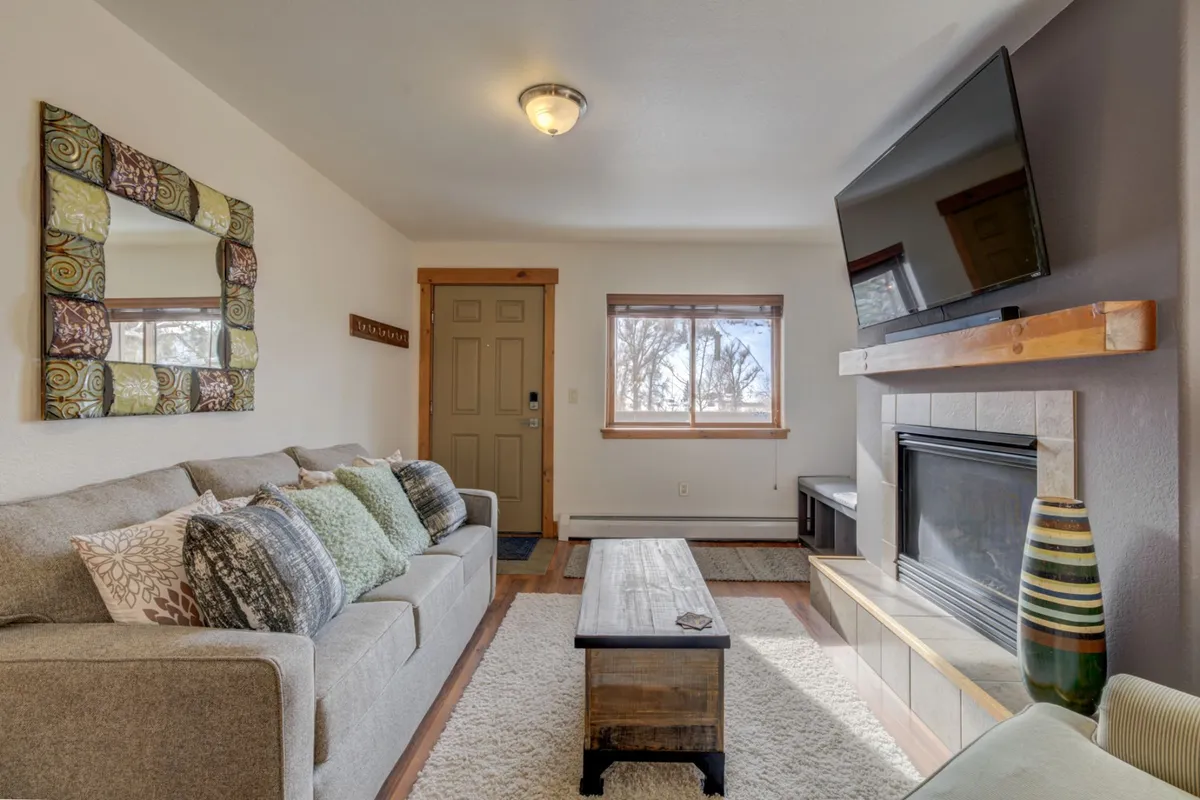 A plush, comfortable sofa with decorative pillows sits in front of a glowing gas fireplace and a flat-screen TV, framed by a wooden coffee table and a large mirror above the mantle. Light streams through a window, inviting guests to relax and enjoy views or entertainment after a day of adventure.